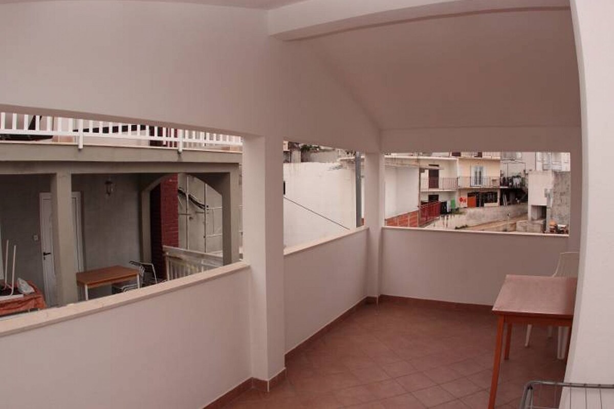 A narrow corridor with a small table and a chair, a white railing, and a view of neighboring buildings.