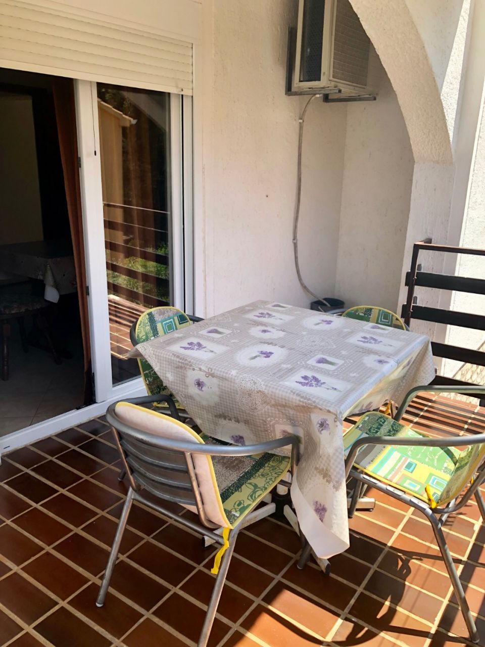 Pequeno alpendre externo com mesa e cadeiras, porta de correr de vidro e ar-condicionado fixado na parede.