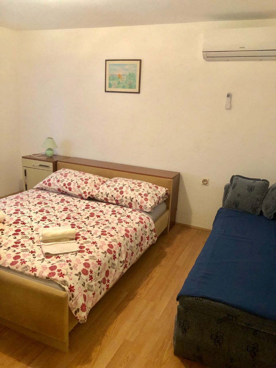 Ein Schlafzimmer mit einem Bett, einem Nachttisch, einer Lampe, einem Bild an der Wand und einer Klimaanlage.