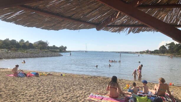 A beach with a thatched roof structure, people sunbathing and swimming, and a calm sea with boats in the distance. - Apartments Kate (Nin)