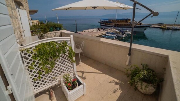 A balcony with a white lattice chair, potted plants, and a view of a marina with boats and a large umbrella. - Apartments Ela (Sutivan)