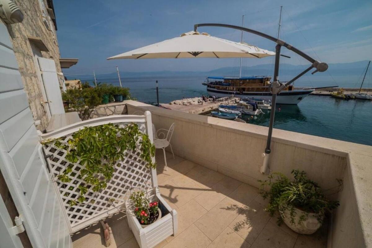 A balcony with a white lattice chair, potted plants, and a view of a marina with boats and a large umbrella.