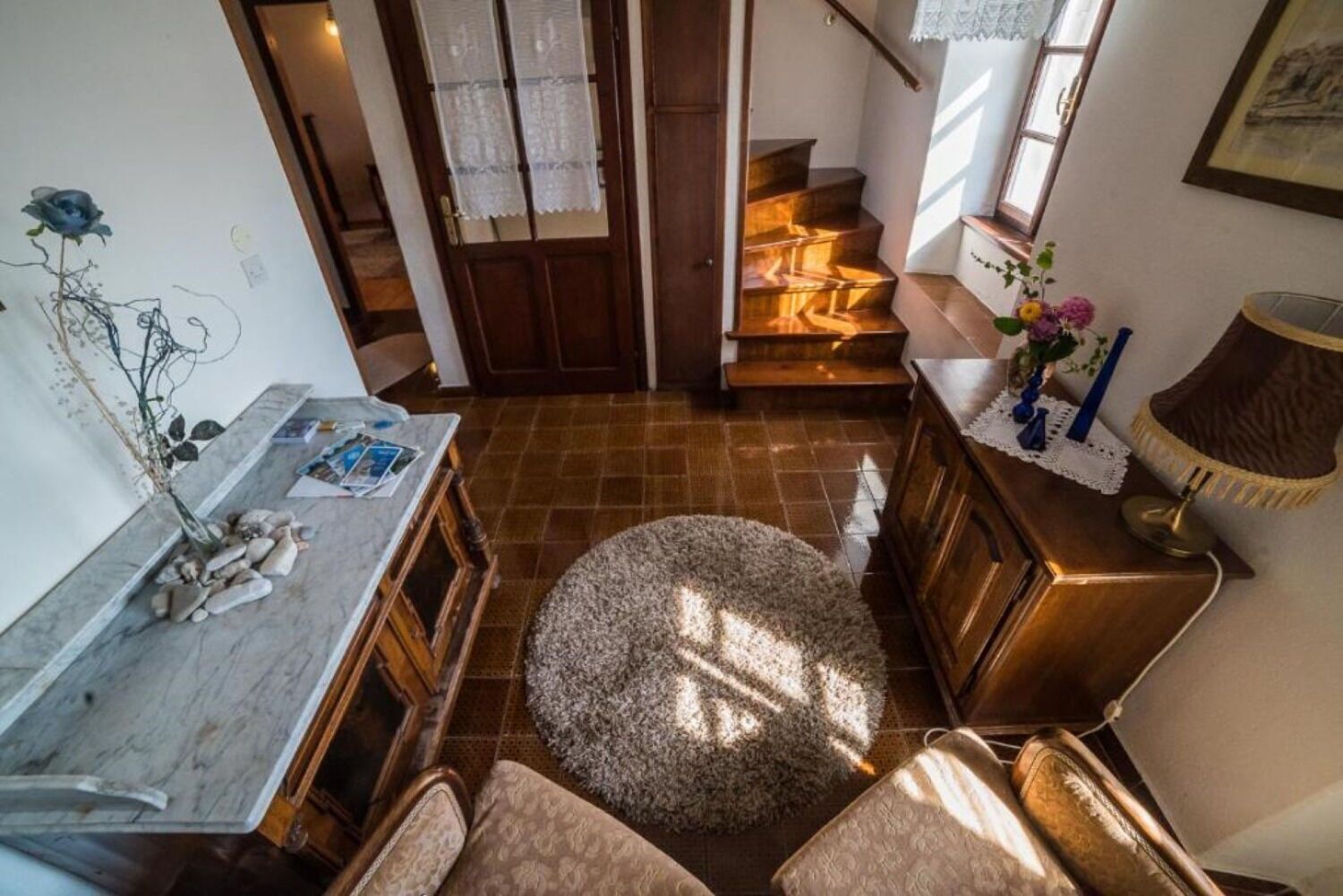 Un salon chaleureux avec une table basse en bois, un tapis rond et un canapé. Un escalier mène à un autre niveau, et il y a une fenêtre ornée de rideaux en dentelle.