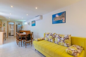A bright yellow sofa with floral cushions, a wooden dining table, and a kitchen area with white cabinets and a refrigerator. - Apartment Mici 1 (Cres)