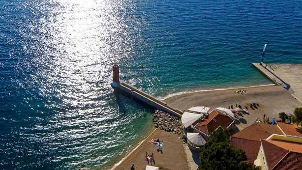 Una zona costiera con un molo che si estende verso il mare, una spiaggia sabbiosa e alcuni edifici con tetti in tegole rosse.