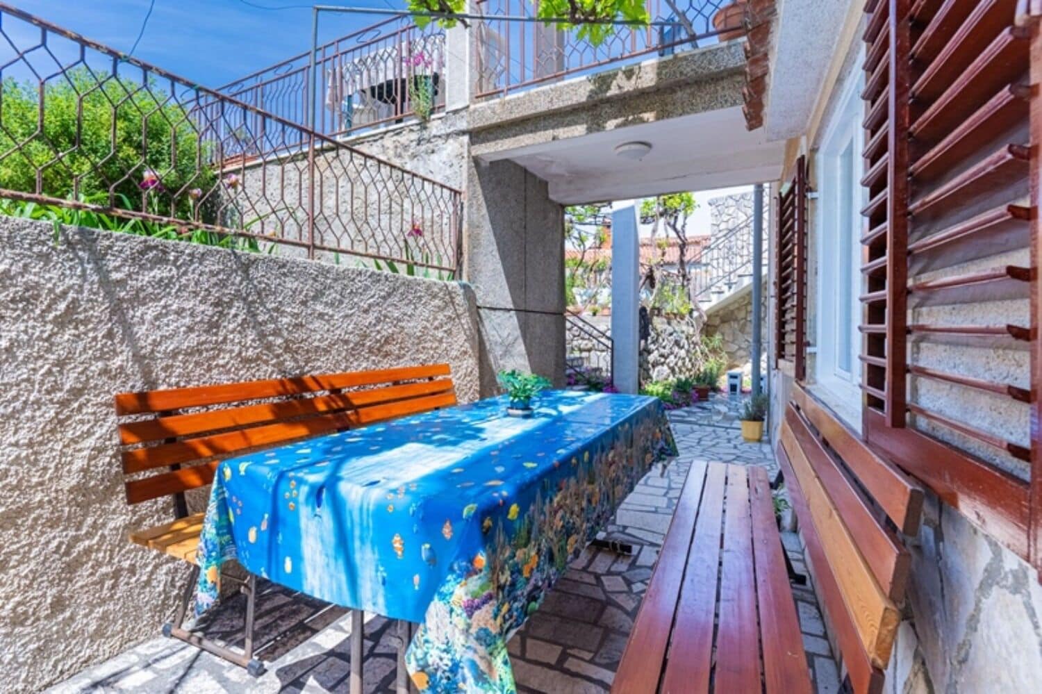 A patio with a wooden bench, a table covered with a blue tablecloth, and a stone wall.