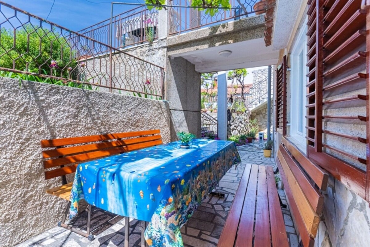 A patio with a wooden bench, a table covered with a blue tablecloth, and a stone wall.