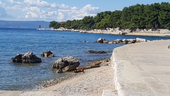 A coastal scene with a pebble beach, large rocks in the water, a stone pathway, and a forested hillside in the background. - Apartment Mici 1 (Cres)