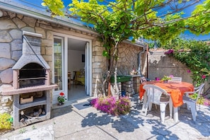 A stone house with a built-in outdoor grill, a dining table with chairs, and a garden with flowers and a tree. - Apartment Mici 1 (Cres)