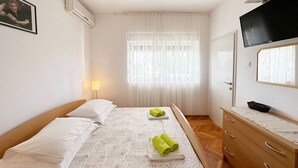 A hotel room with a bed, a bedside table with a mirror, a TV mounted above the table, and a window with sheer curtains. - Apartments Tereza (Trogir)