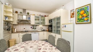 A compact kitchen with white cabinets, a microwave, and a refrigerator. There is a dining table with a floral tablecloth and a bowl in the center. - Apartments Tereza (Trogir)