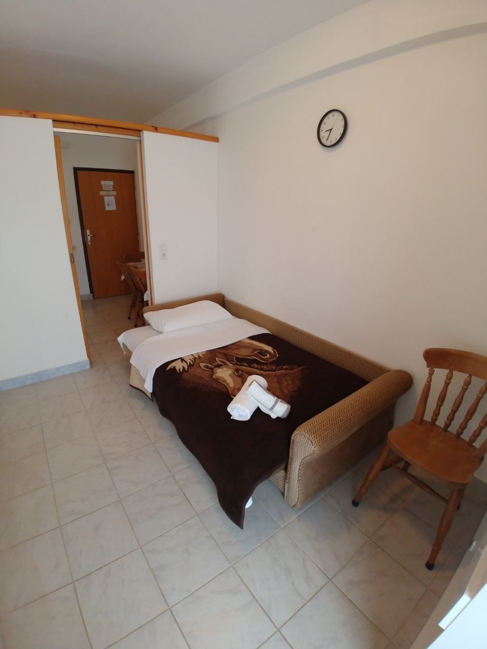 Une petite pièce, bien rangée, avec un lit, une chaise et une horloge au mur.
