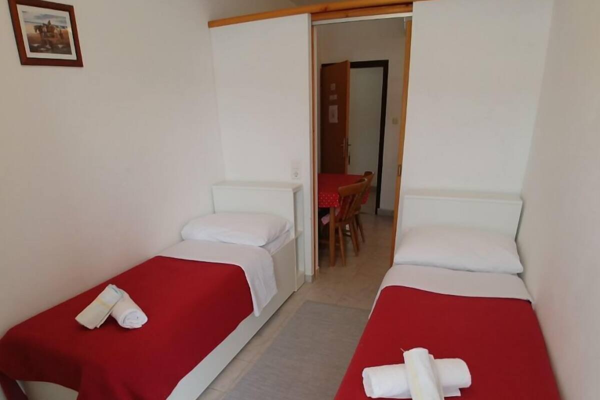 Une chambre avec deux lits simples, chacun recouvert d’une couverture rouge et agrémenté de serviettes pliées. On aperçoit une petite table recouverte d’une nappe rouge, ainsi que des chaises à l’arrière-plan.