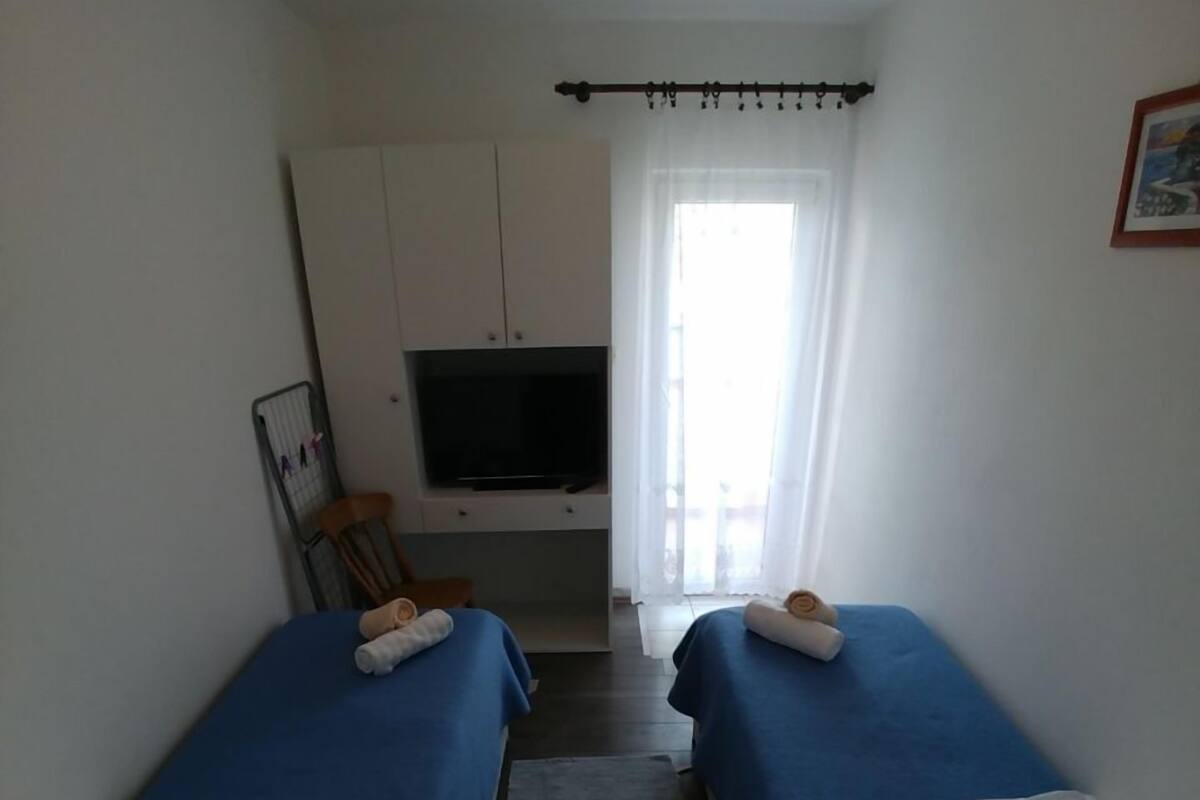 Une petite chambre d’hôtel étroite, avec deux lits simples, une télévision, une chaise et une fenêtre avec des rideaux.