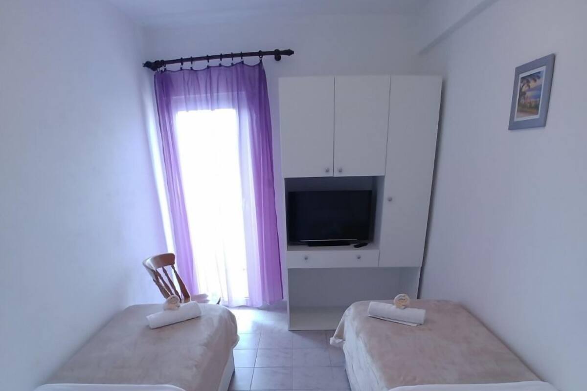 Une petite chambre propre, avec deux lits, une télévision et une fenêtre ornée de rideaux violets.