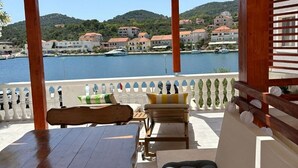 Eine Holzterrasse mit Tisch und Stühlen, einer gepolsterten Bank und Blick auf ein Gewässer sowie Gebäude im Hintergrund.
