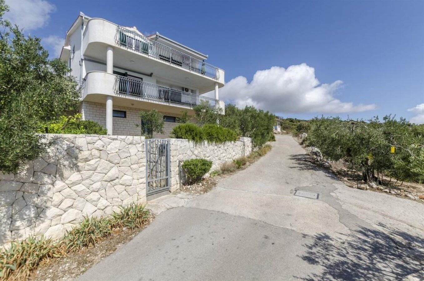 Un edificio residencial de dos plantas con balcones, una pared de piedra y un camino pavimentado que conduce a un viñedo.