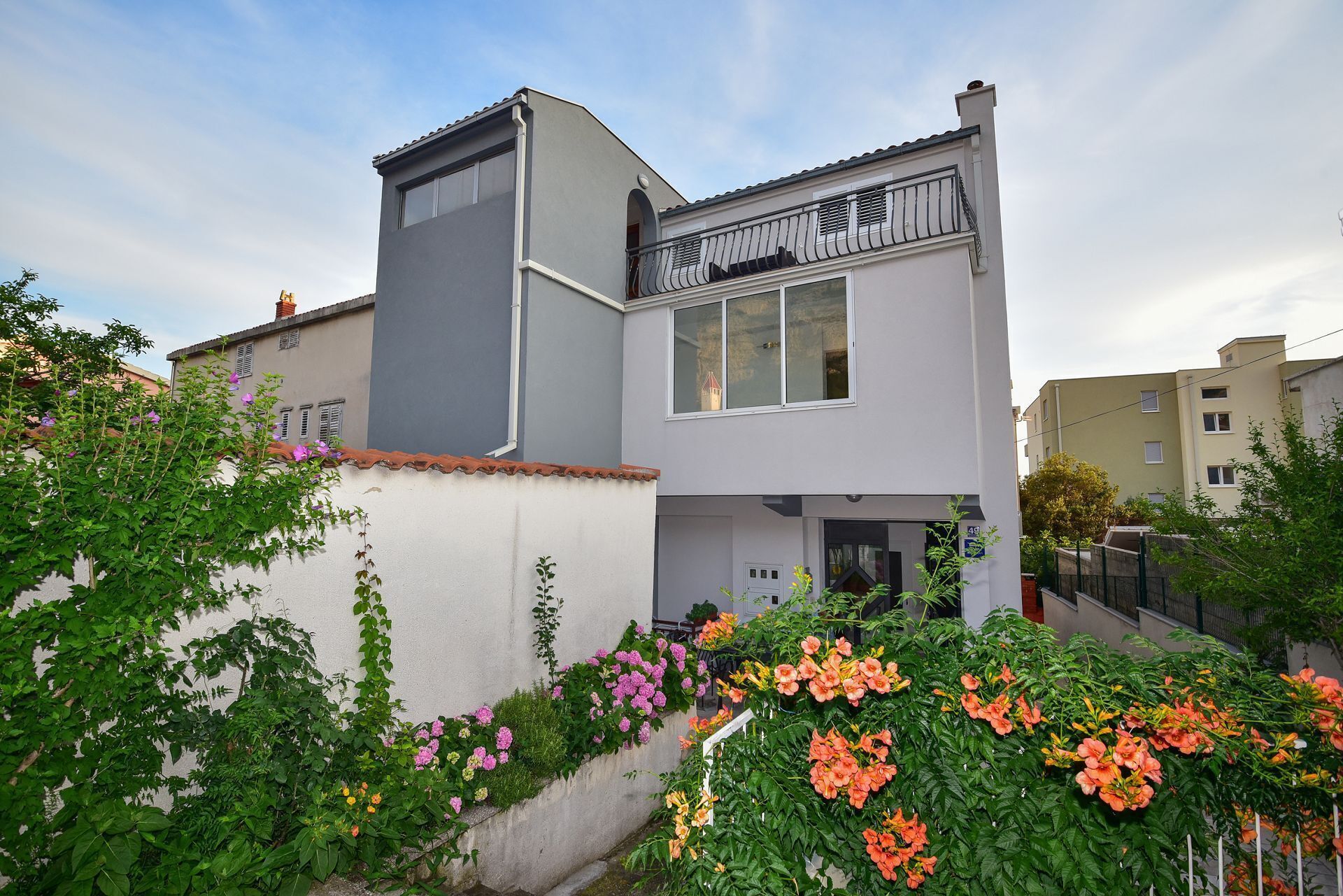 A modern two-story house with a balcony, surrounded by lush greenery and flowering plants.