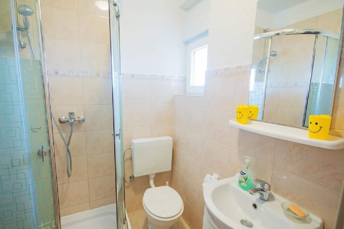 Un bagno con box doccia in vetro, un water, un lavabo con dispenser di sapone e uno specchio con ripiano contenente vasetti gialli a forma di faccine sorridenti.
