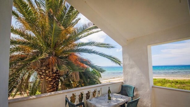 A balcony with a table and chairs, overlooking a beach and palm tree. - Apartments Teo (Privlaka)