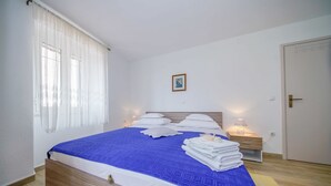 A neatly made bed with a blue and white bedspread, white pillows, and a stack of white towels on the floor beside it. There is a bedside table with a lamp, a framed picture on the wall, and a door to the right. - Apartment Nedjeljka (Postira)