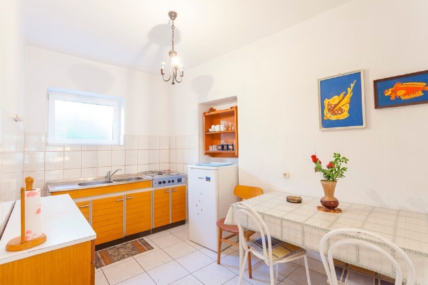 Een kleine keuken met een spoelbak, kookplaat en een koelkast, een eettafel met stoelen, en een kroonluchter.