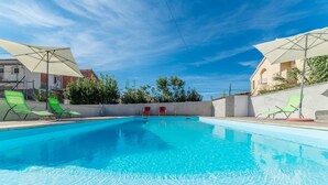 Una piscina azul cristalina con sillones de descanso verdes y rojos, rodeada de sombrillas y un muro bajo con árboles.