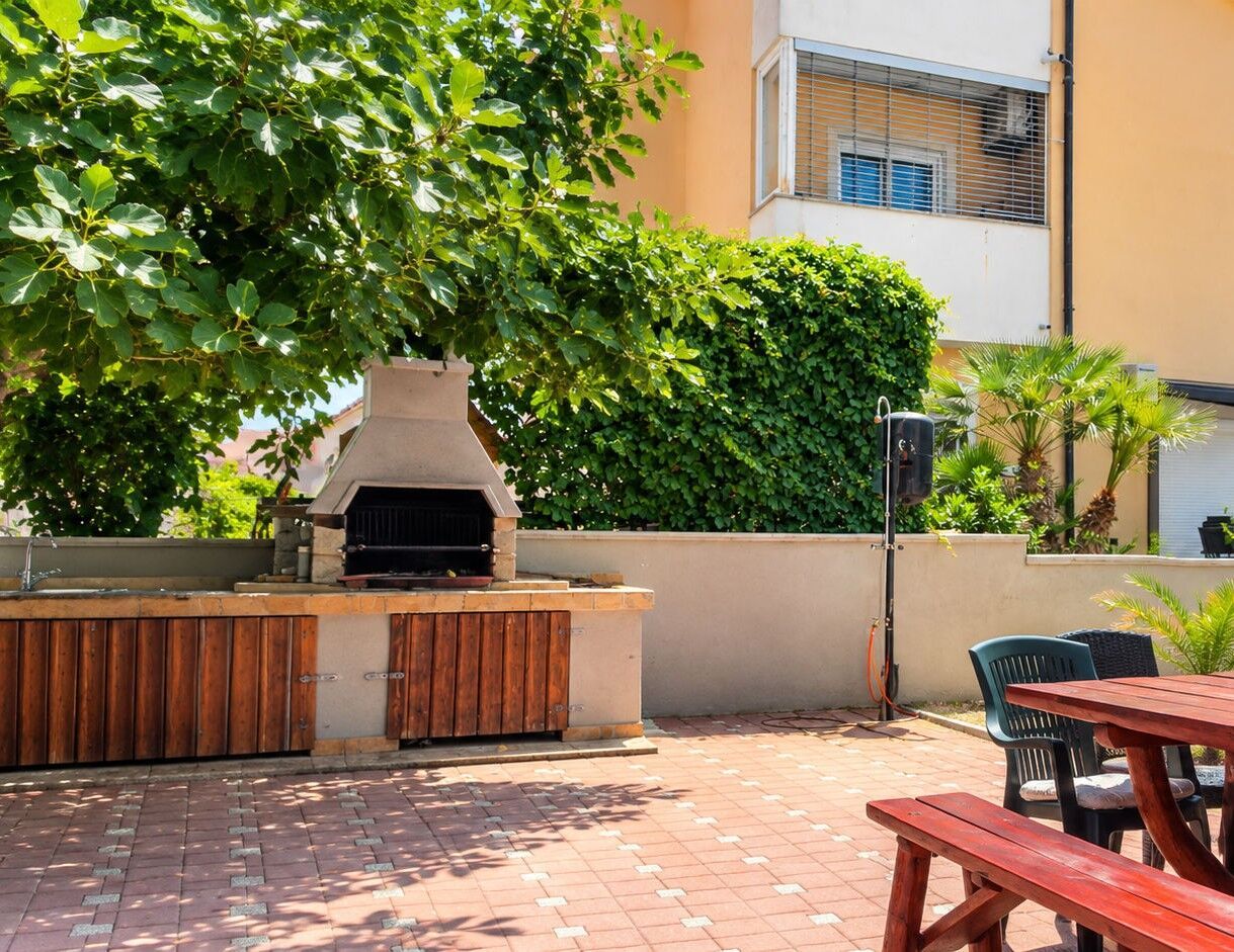 An outdoor barbecue area with a built-in grill, wooden seating, and a paved patio.