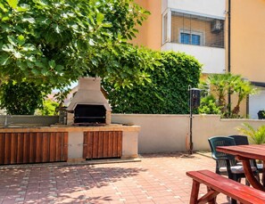 An outdoor barbecue area with a built-in grill, wooden seating, and a paved patio. - Apartments Sab (Povljana)