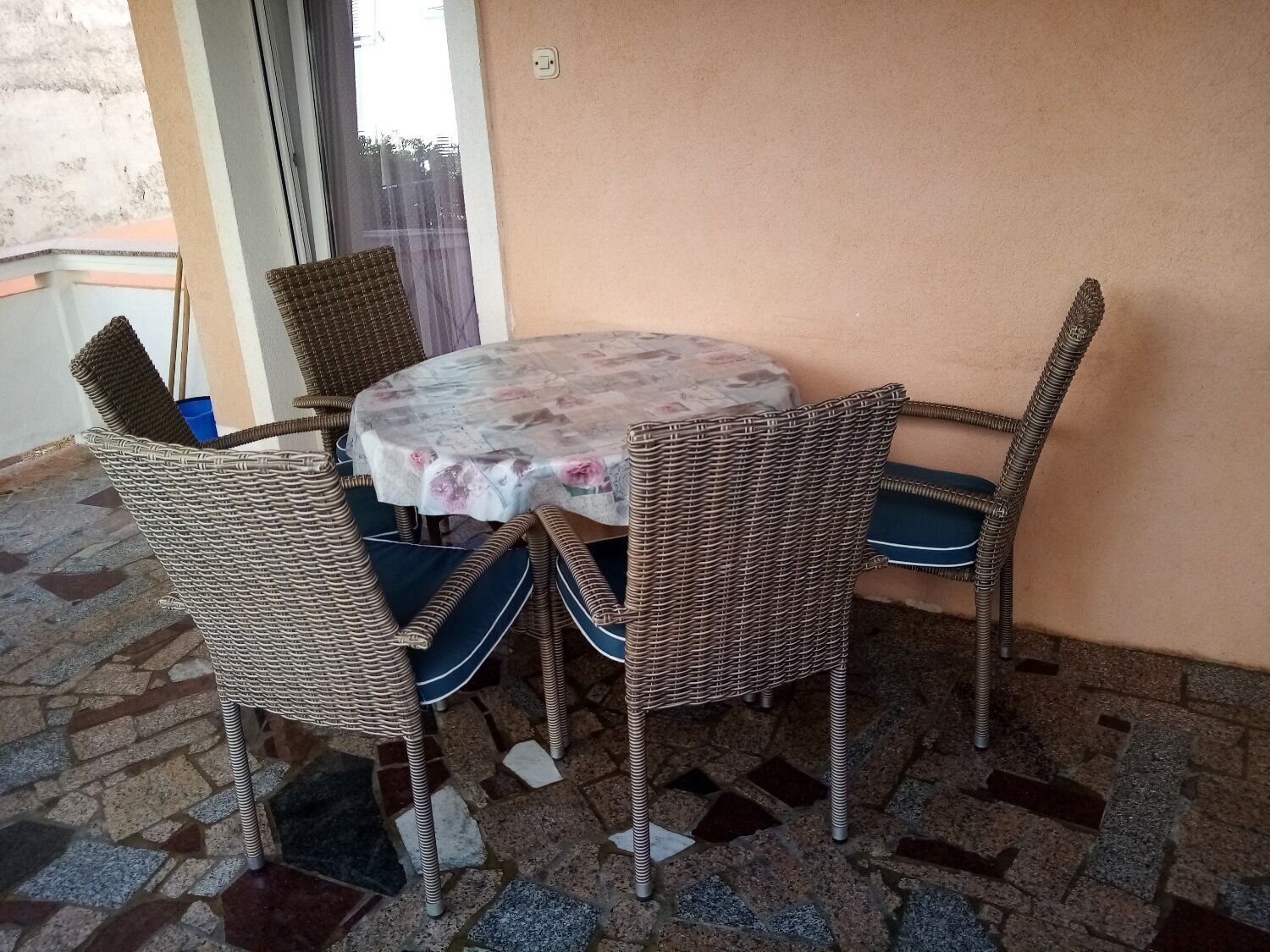 A round table with a floral tablecloth surrounded by wicker chairs with blue cushions.