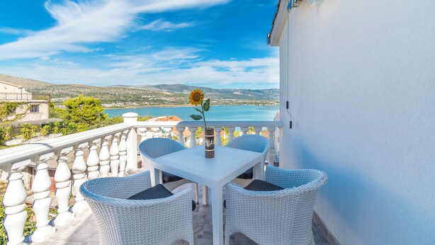 Ein Balkon mit einem weißen Tisch und Korbstühlen, der auf eine Küstenlandschaft blickt.
