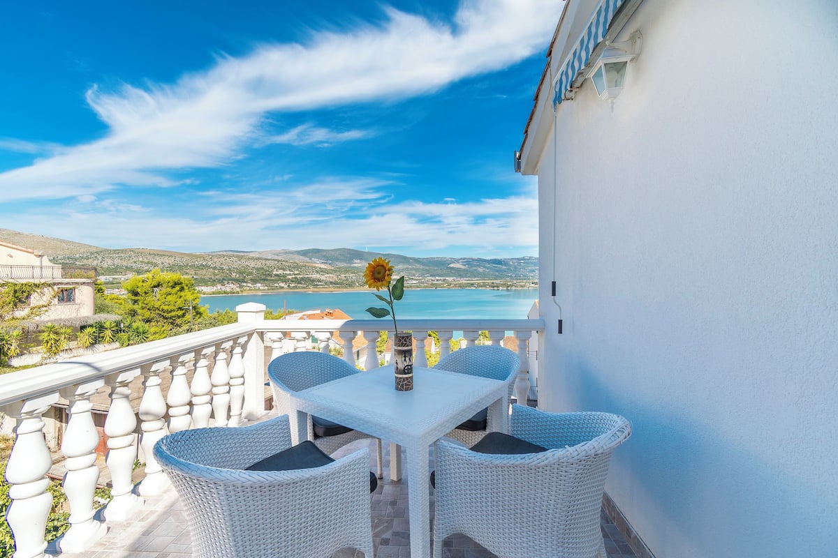 A balcony with a white table and wicker chairs, overlooking a coastal landscape.