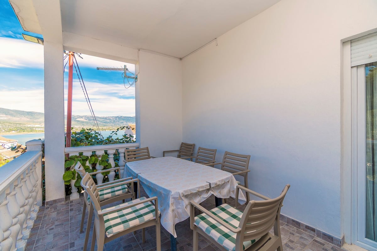 A balcony with a table and chairs, a view of the sea, and a window with a curtain.