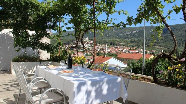An outdoor dining area with a table set for two, overlooking a town with red-roofed buildings and a mountainous backdrop. - Apartment Jasna (Jelsa)