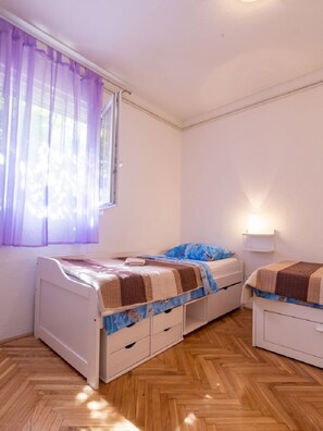 A room with two beds, a window with purple curtains, a bedside lamp, and a herringbone wooden floor. - Apartments Petar (Trogir)