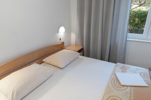 Een net opgemaakt bed met witte lakens en kussens, een nachtkastje met een lamp, en een raam met gordijnen.