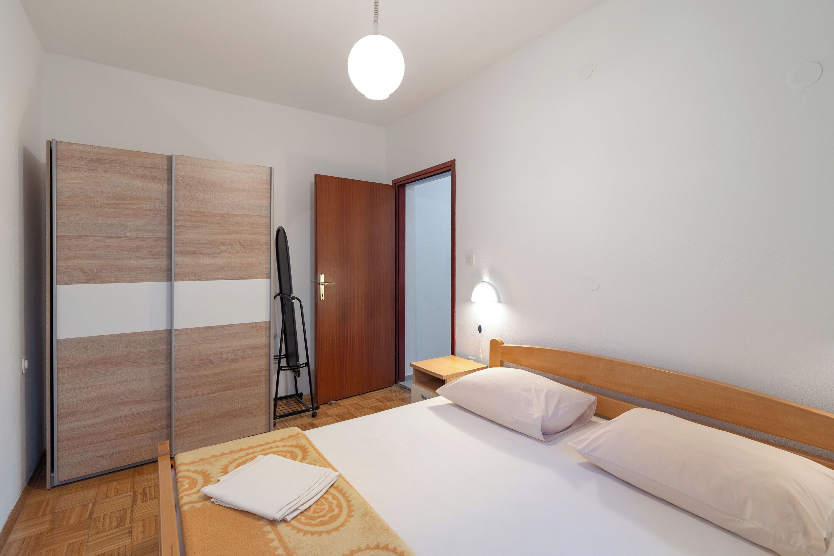 A hotel room with a bed, a wooden wardrobe, a desk with a chair, and a wall-mounted light.