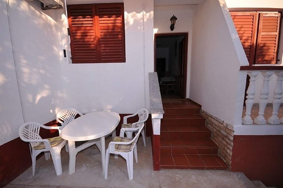 Eine kleine Außenpatio mit einem runden Tisch und vier Stühlen, eine Treppe, die zu einer oberen Ebene führt, und Holzläden an den Wänden.