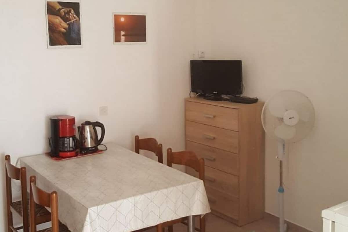Ein Essbereich mit Tisch, Stühlen, einer Kaffeemaschine, einem Fernseher auf einer Kommode und einem Standventilator.