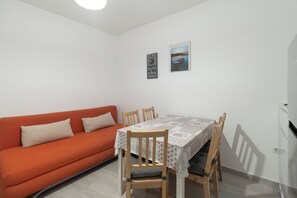Een lichte, kleine kamer met een okergele bank, een houten eettafel met stoelen, en een witte muur met twee ingelijste foto’s.