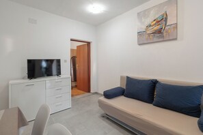 A room with a flat-screen TV on a white cabinet, a beige sofa with blue cushions, and a painting on the wall. - Apartments Pere (Povljana)