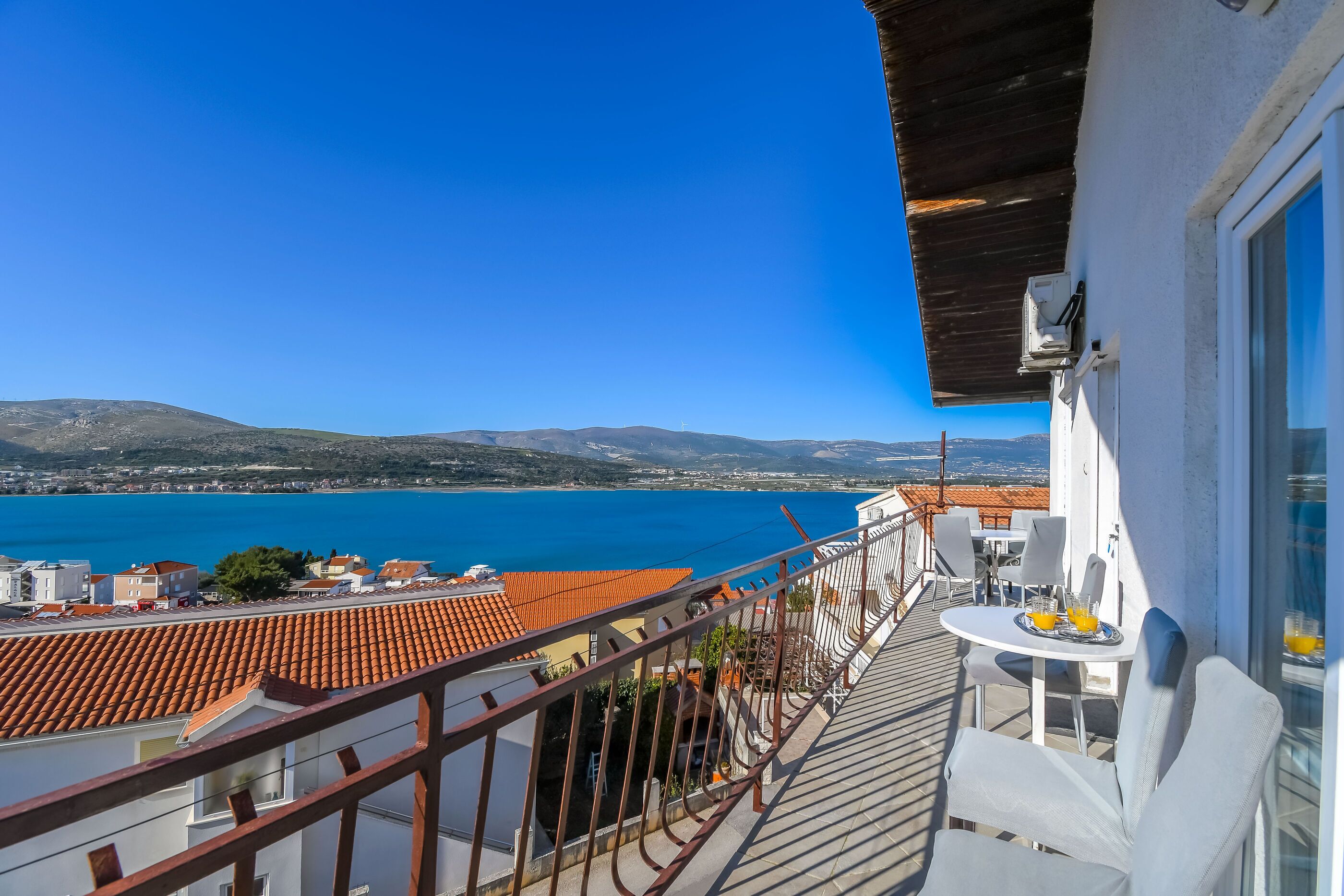 A balcony with a view of the sea and distant hills, a table with chairs, and a small table with a tray of drinks.