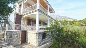 Un bâtiment en pierre de deux étages, doté d’un balcon, entouré de verdure et avec une chaîne de montagnes en arrière-plan.