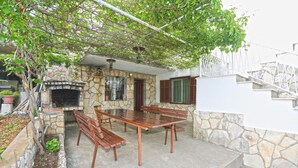 Une terrasse extérieure revêtue de pierres, avec une table et des bancs en bois, une cheminée en pierre et un escalier menant à un niveau supérieur.