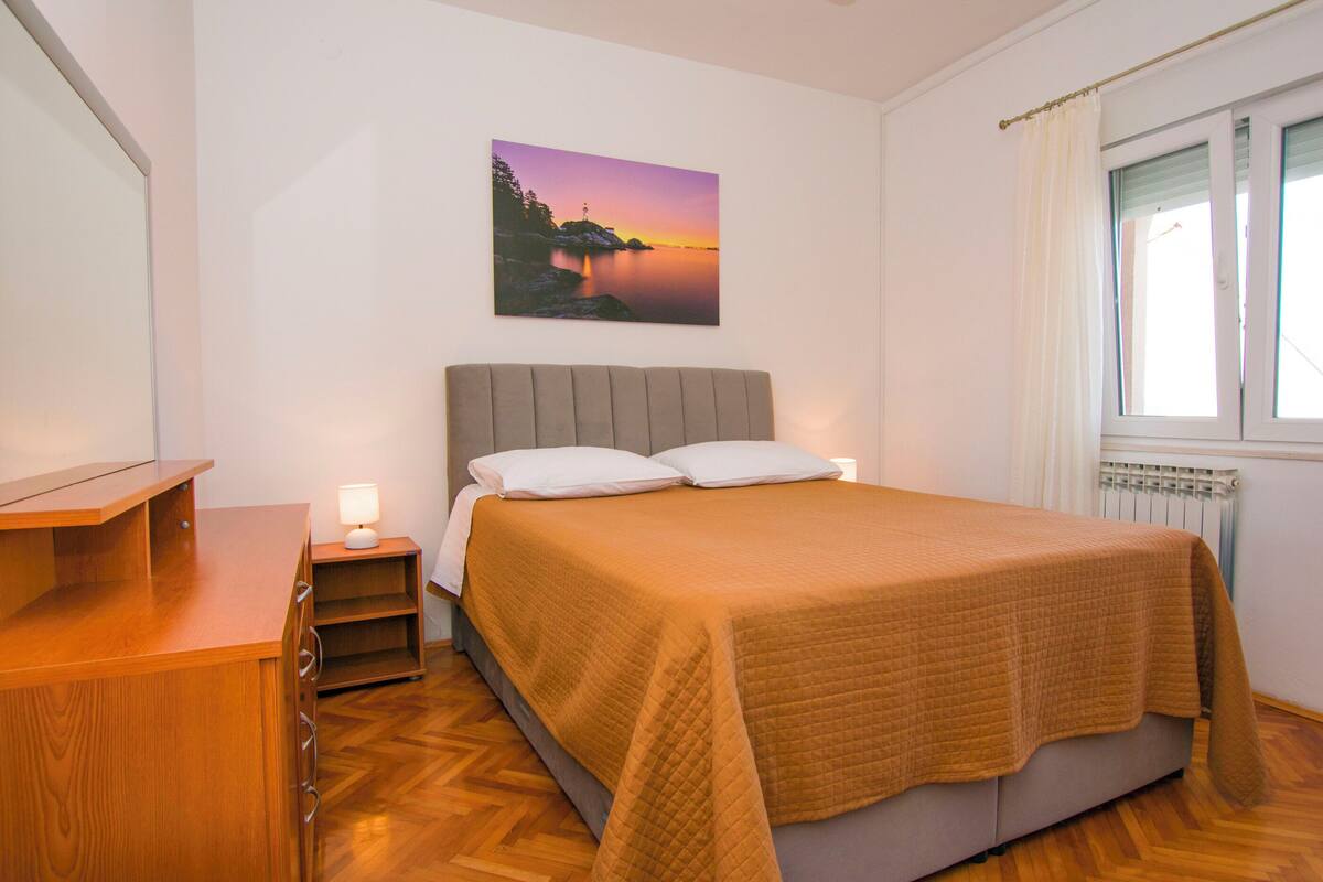 Une chambre à coucher avec un lit, une table de chevet, une commode en bois, une fenêtre avec des rideaux et un tableau accroché au mur.