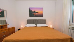 Ein Schlafzimmer mit einem Bett, zwei Nachttischlampen, einem Nachttisch, einer Kommode, einem Spiegel und einem Bild von einem Sonnenuntergang über Wasser an der Wand.