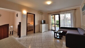 Una sala de estar con un sofá, una mesita de centro, un televisor y una pequeña mesa. Hay una puerta que da a otra habitación.