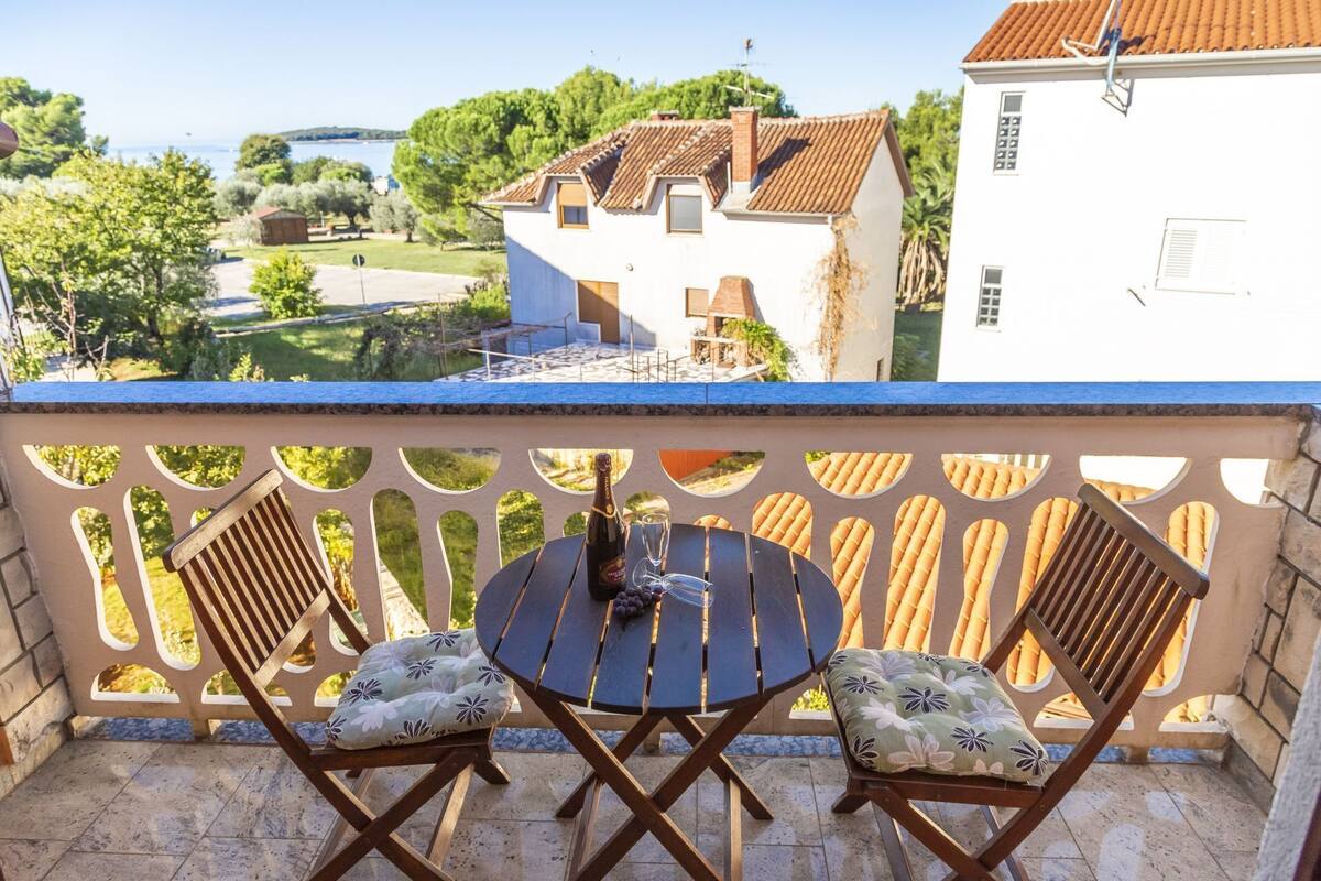 Un balcon avec deux chaises en bois et une petite table ronde, donnant sur un paysage pittoresque avec un bâtiment blanc et des arbres.