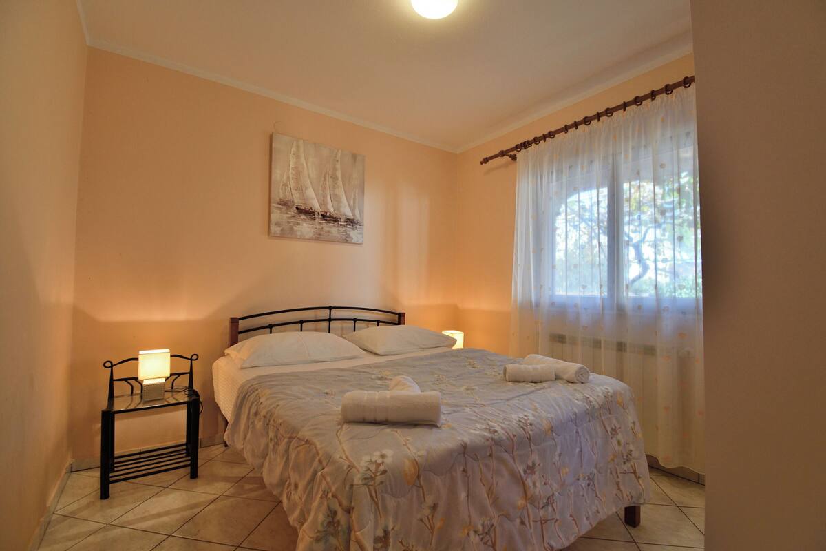 Une chambre à coucher avec un lit, une table de chevet, une lampe, un tableau au mur et une fenêtre avec des rideaux.