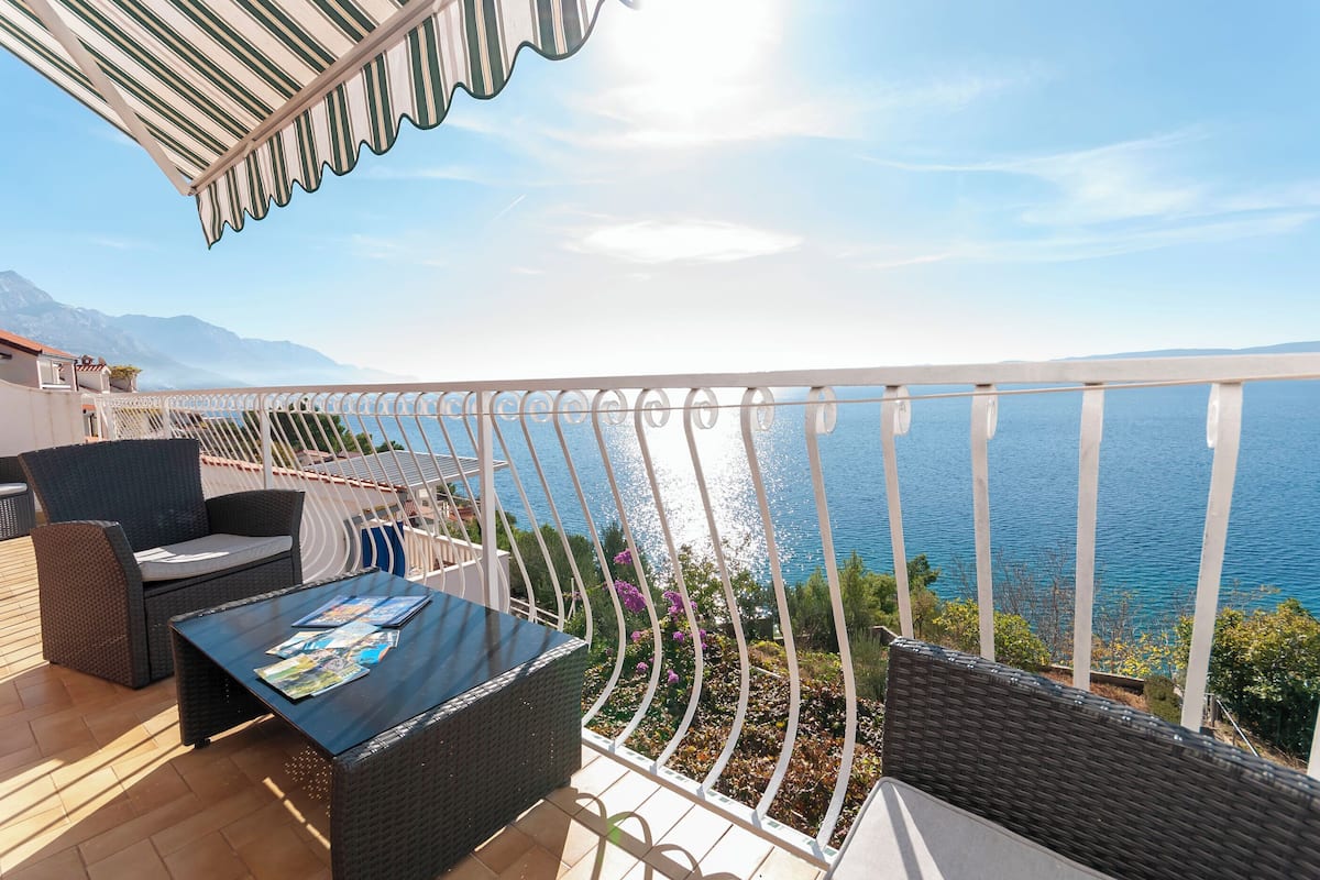 Een balkon met rieten meubels, een tafel met tijdschriften en uitzicht op zee en bergen.